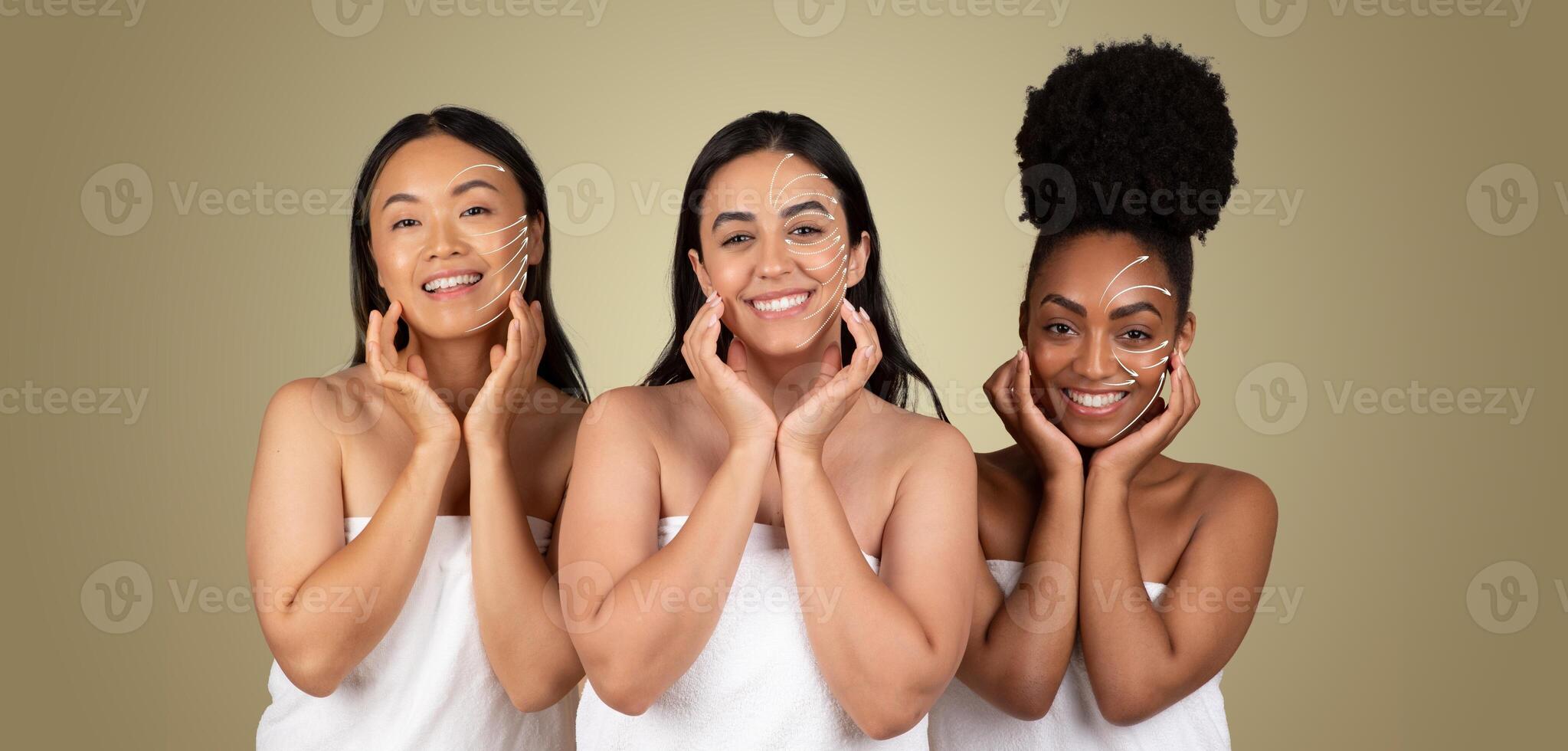 Three diverse women are smiling while receiving facial treatments. They are wrapped in towels, showcasing a relaxed atmosphere focused on skin care and anti-aging techniques in a soothing environment. photo
