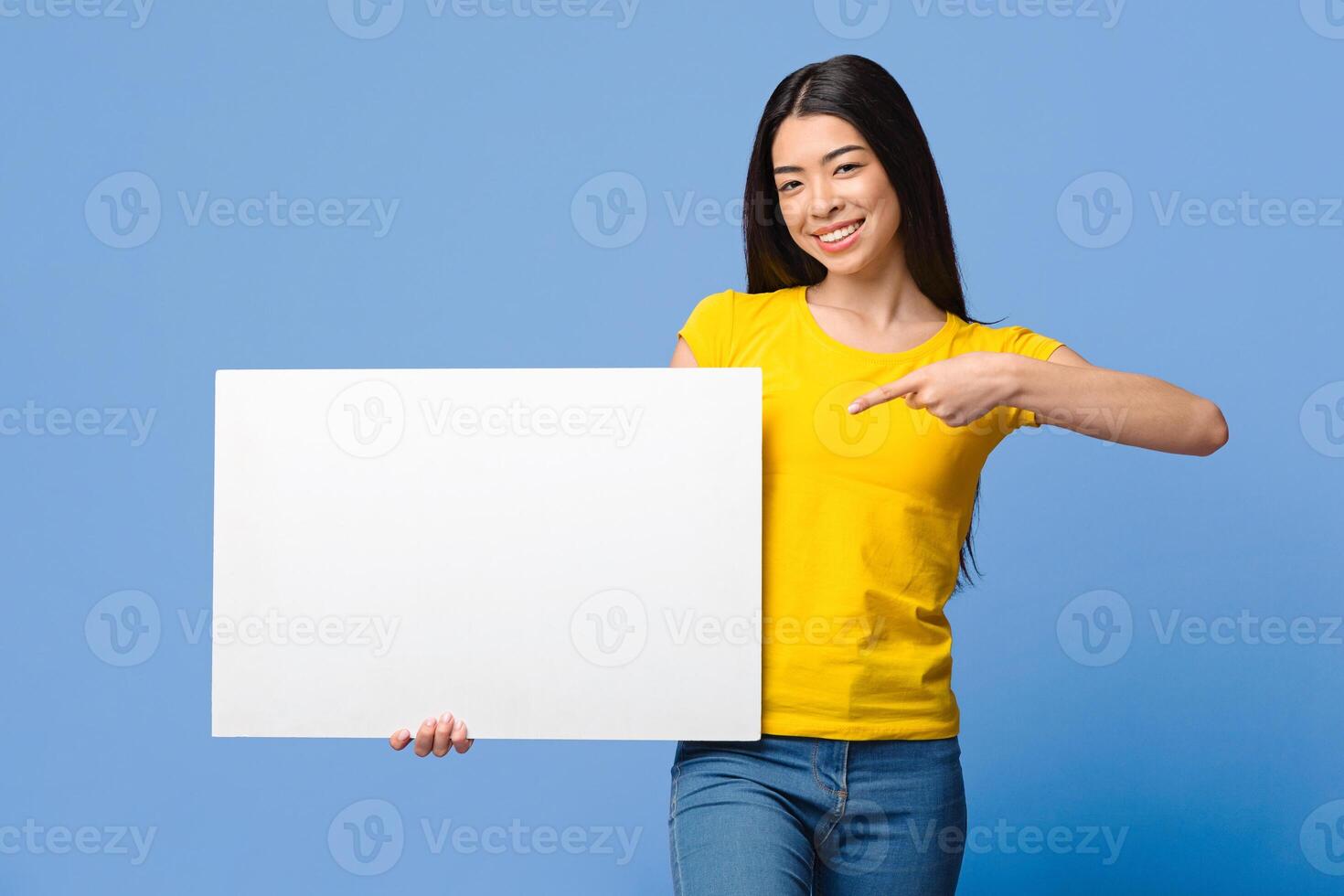 Place For Your Ad. Smiling Asian Girl Holding White Blank Placard, Pointing At Copy Space, Posing Over Purple Studio Background photo