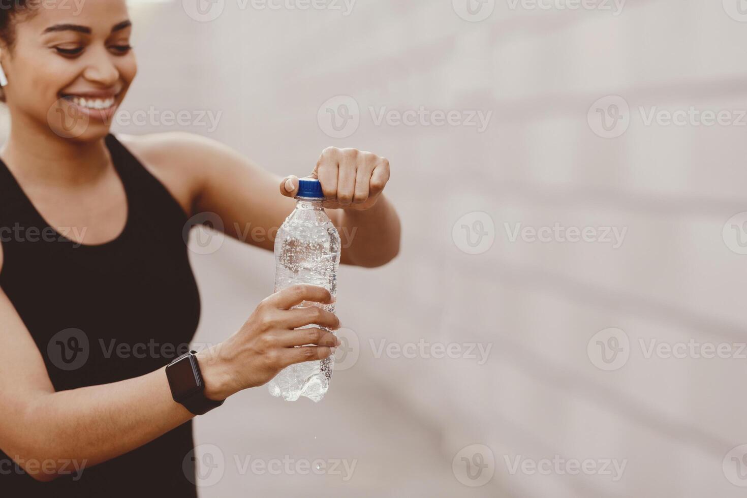 Vital energy, water and sport. Happy african american girl in sportswear with modern headphones and smart watch, opens bottle on training in city, close up, copy space photo