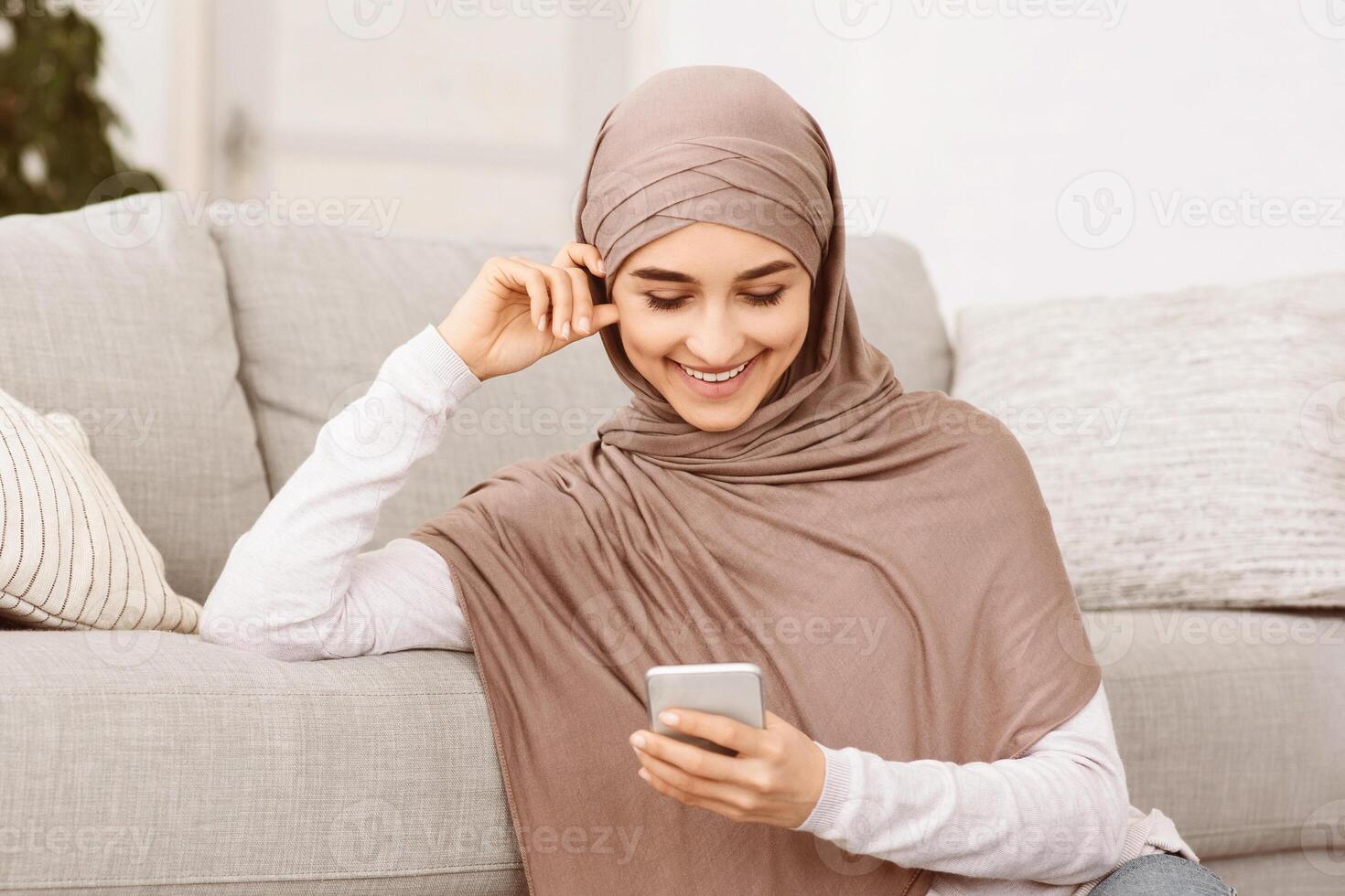 Nice App. Happy Muslim Girl In Hijab Using Smartphone At Home, Checking New Application, Browsing Internet Or Messaging With Friends, Closeup photo
