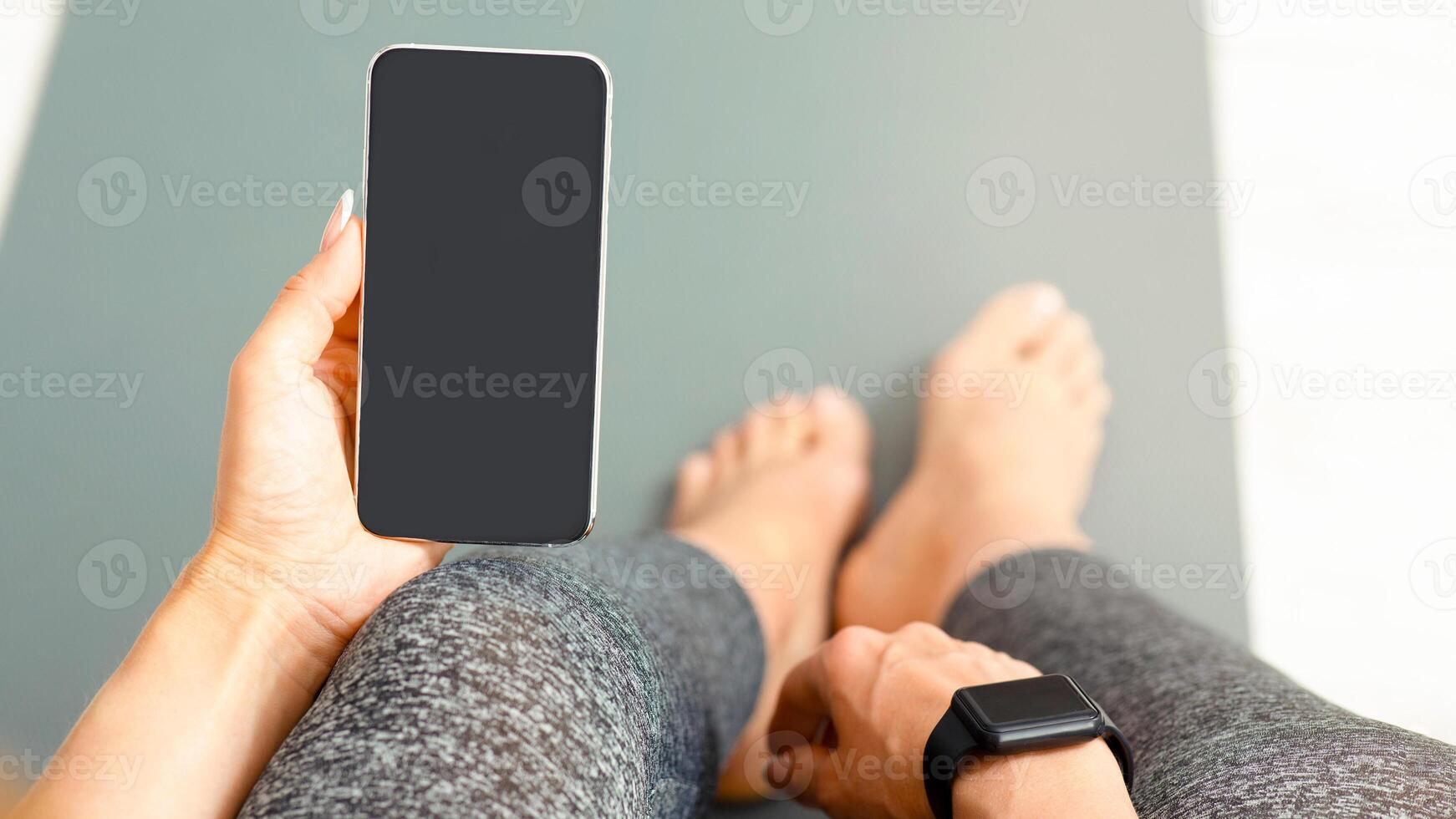 Modern gadgets for training at home. Woman sitting on mat with fitness tracker and smartphone in hand with blank screen, free space photo