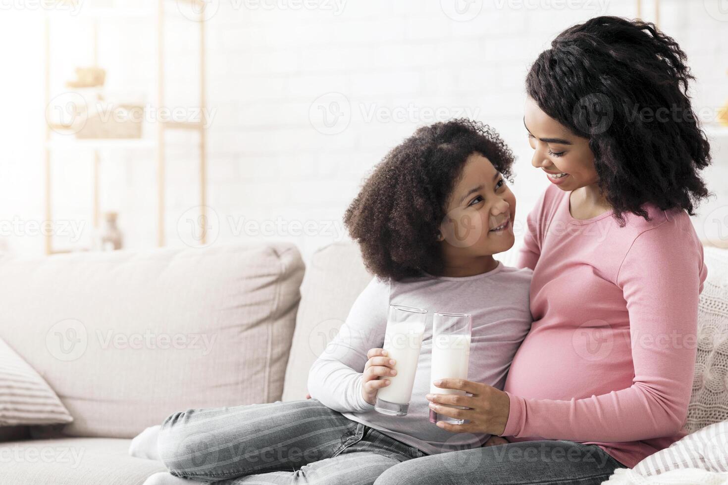 Little african american girl and her pregnant mother drinking milk sitting on sofa at home, clinking glasses, having fun together, free space photo