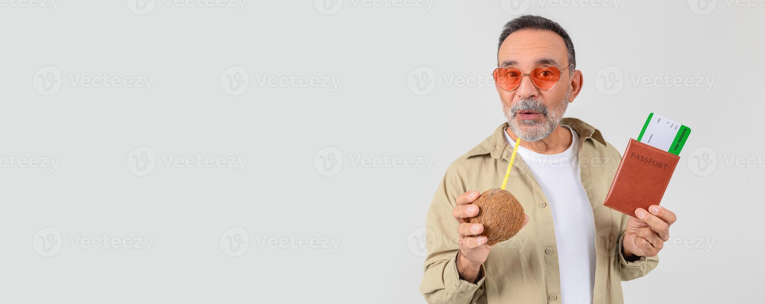 Joyful senior man in funky sunglasses sips from a coconut while holding a passport and boarding pass, suggesting a tropical vacation photo