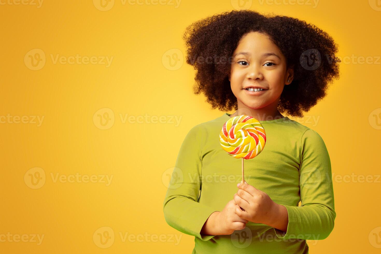 Adorable Little African American Girl Holding Lollipop Candy In Hands, Posing To Camera Over Yellow Background And Smiling, Copy Space photo