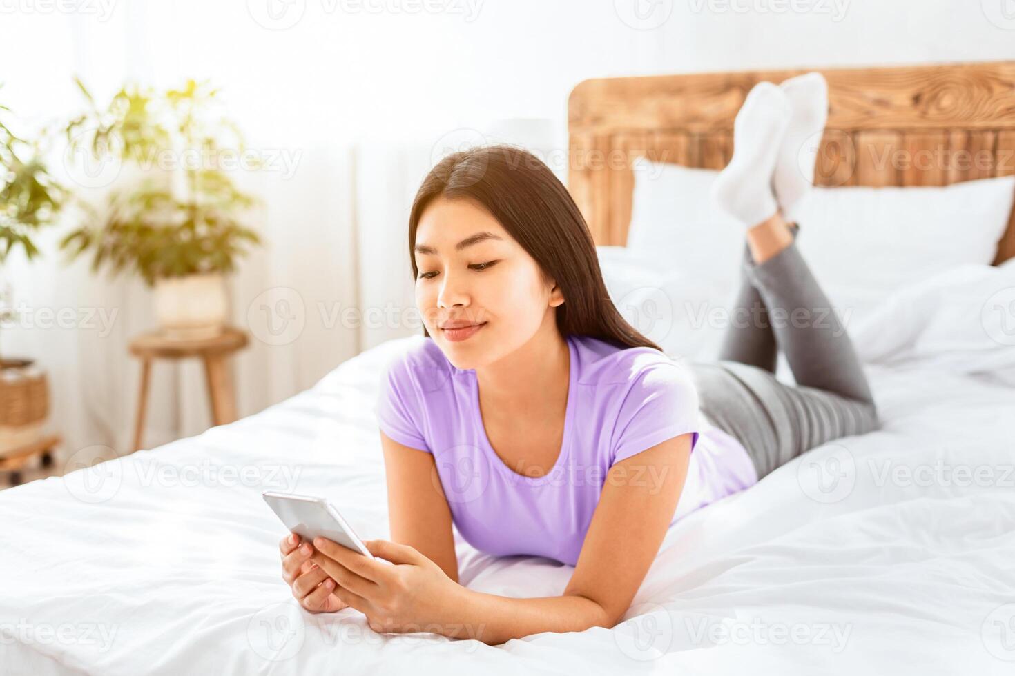 Chinese Woman With Smartphone Using Mobile Application Or Texting Lying On Bed At Home. Modern Communication photo