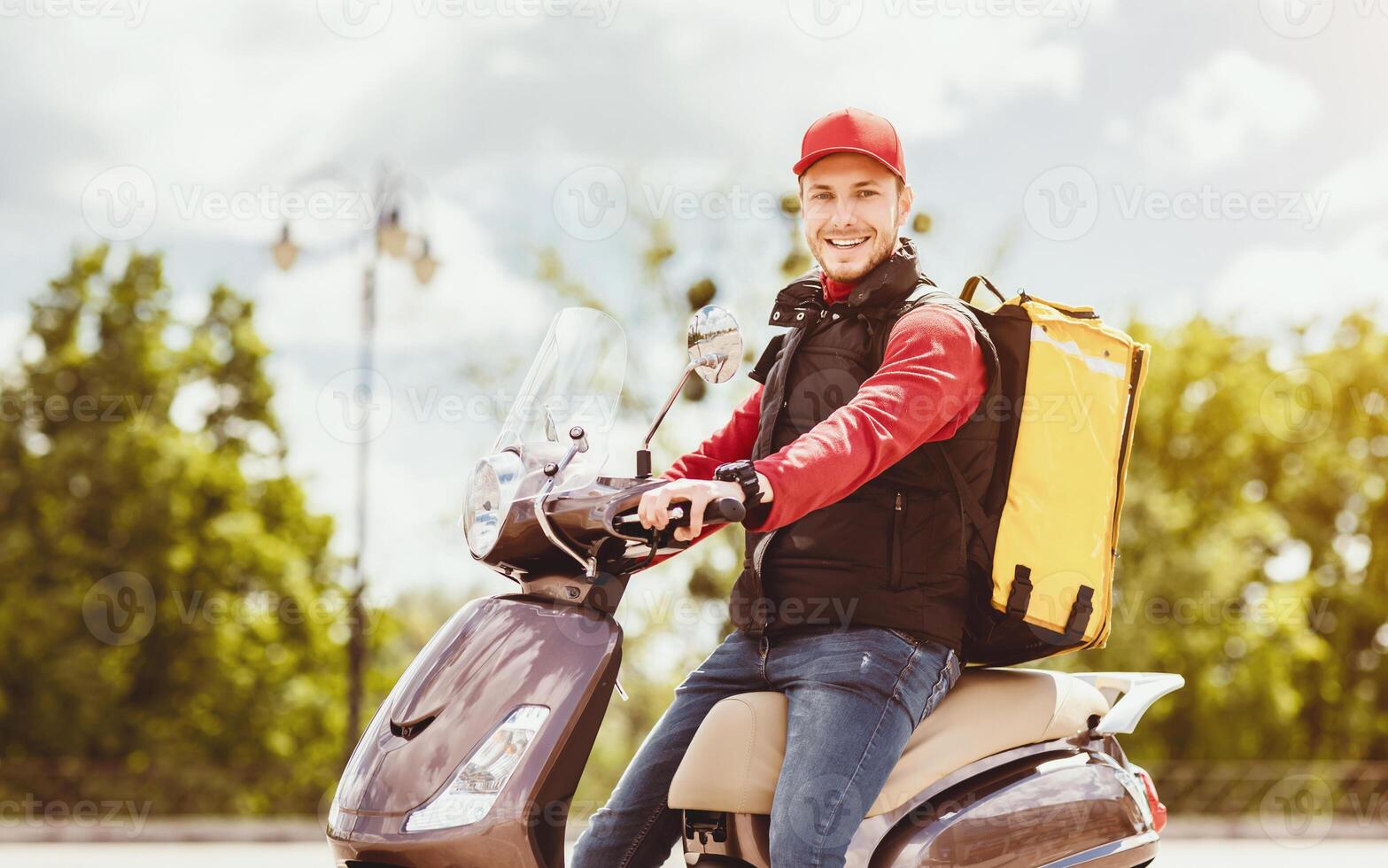 Delivering Food. Courier Man With Yellow Backpack Sitting On Moto Scooter Smiling To Camera Outside. Free Space photo