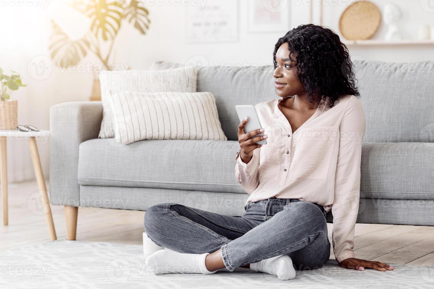 Millennial Black Woman Sitting With Smartphone On Floor At Home, Relaxing In Living Room, Dreamy African American Female Looking Away And Thinking About Something, Enjoying Day Off Free Time photo