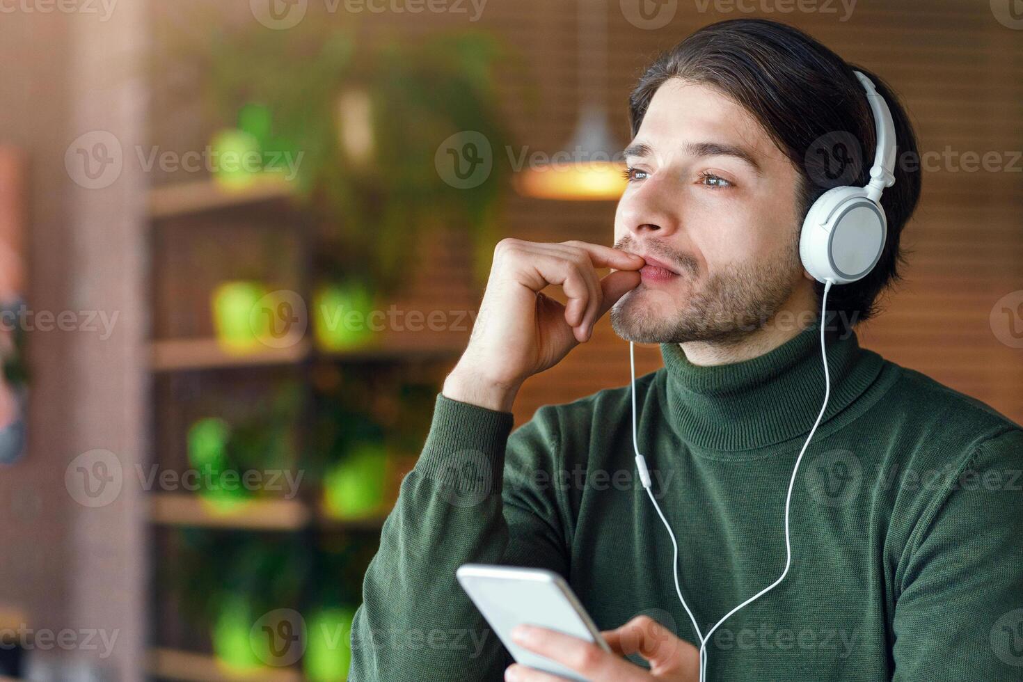 Pensive young man in headphones listening to music, using mobile phone, drinking coffee, cafe interior, empty space photo