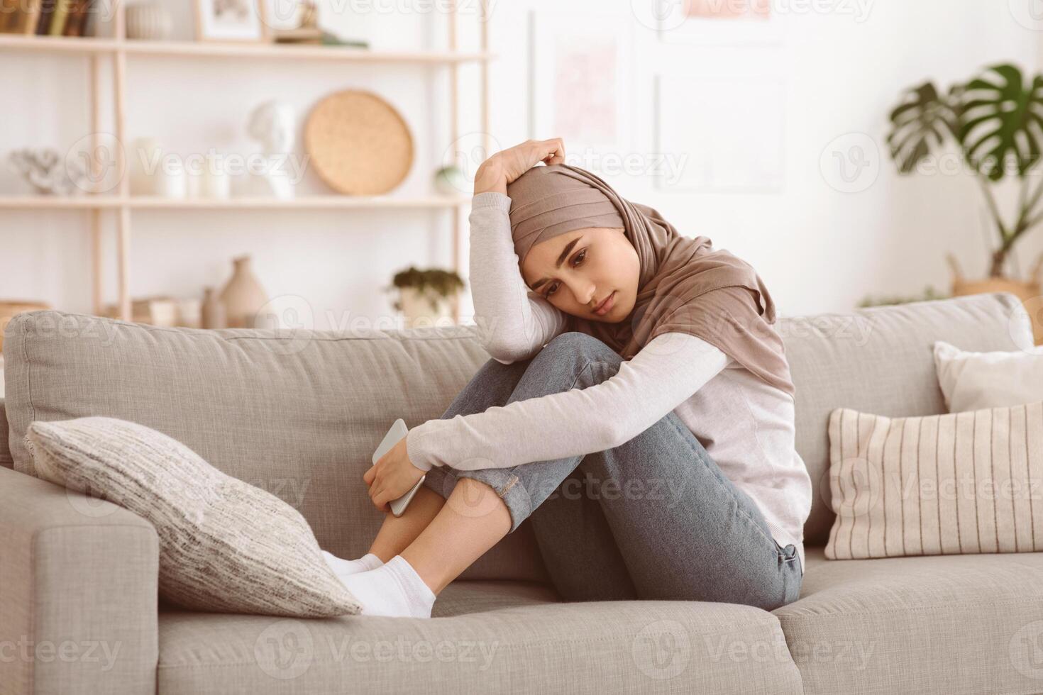 Depressed arabic woman in hijab sitting on couch at home with smartphone in hands, feeling lonely, looking away with sad face expression, copy space photo