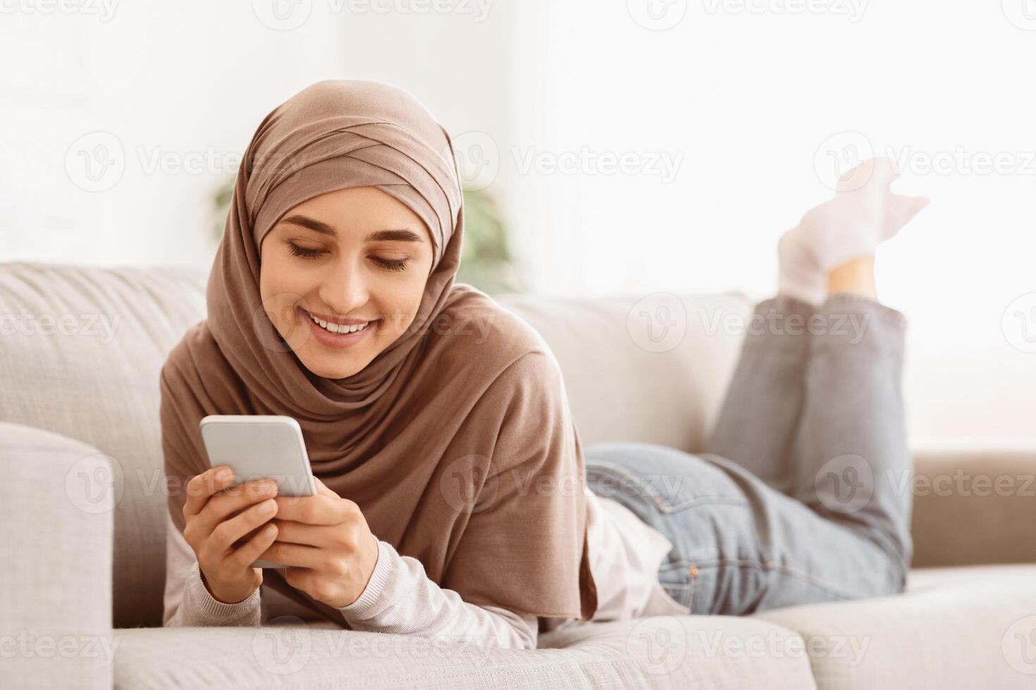 Beautiful arabic girl in hijab texting on smartphone while lying on cozy couch at home, chatting with friends or browsing social networks, closeup photo