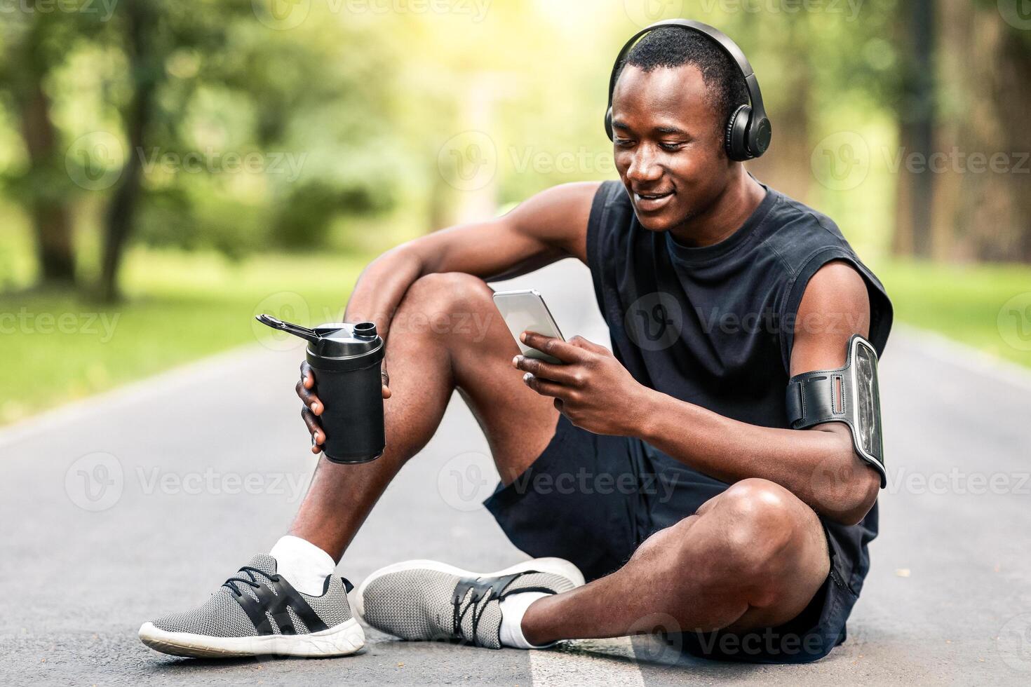 Morning exercising. Happy athletic black guy resting on path at public park, using phone and drinking water, copy space photo