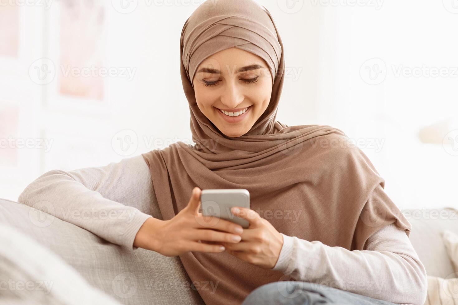 Social Networks. Smiling arab woman in hijab messaging on smartphone at home, relaxing on comfortable couch, closeup shot with copy space photo