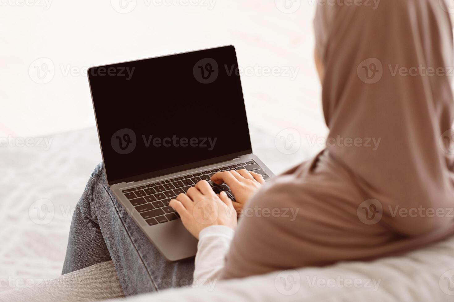 Mockup image of unrecognizable muslim woman working on laptop with black screen at home, sitting on couch, over shoulder view with free space photo