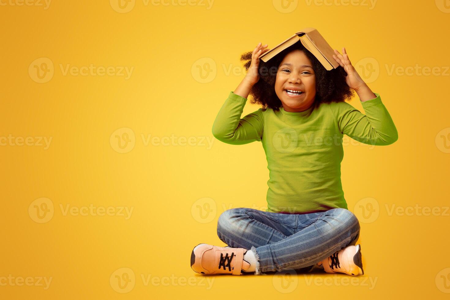 Education Concept. Portrait Of Playful Little Afro Girl With Opened Book On Head Sitting On Yellow Background In Studio And Laughing, Copy Space photo