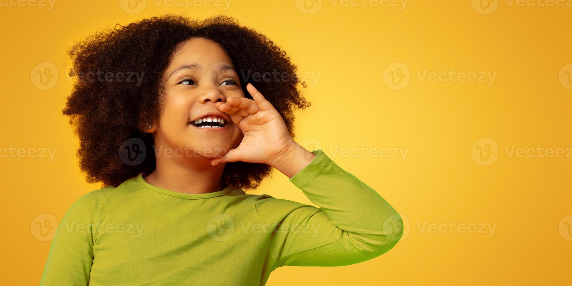 Overjoyed cute little black child holding hand near mouth, making announcement, screaming aside at copy space on yellow background photo