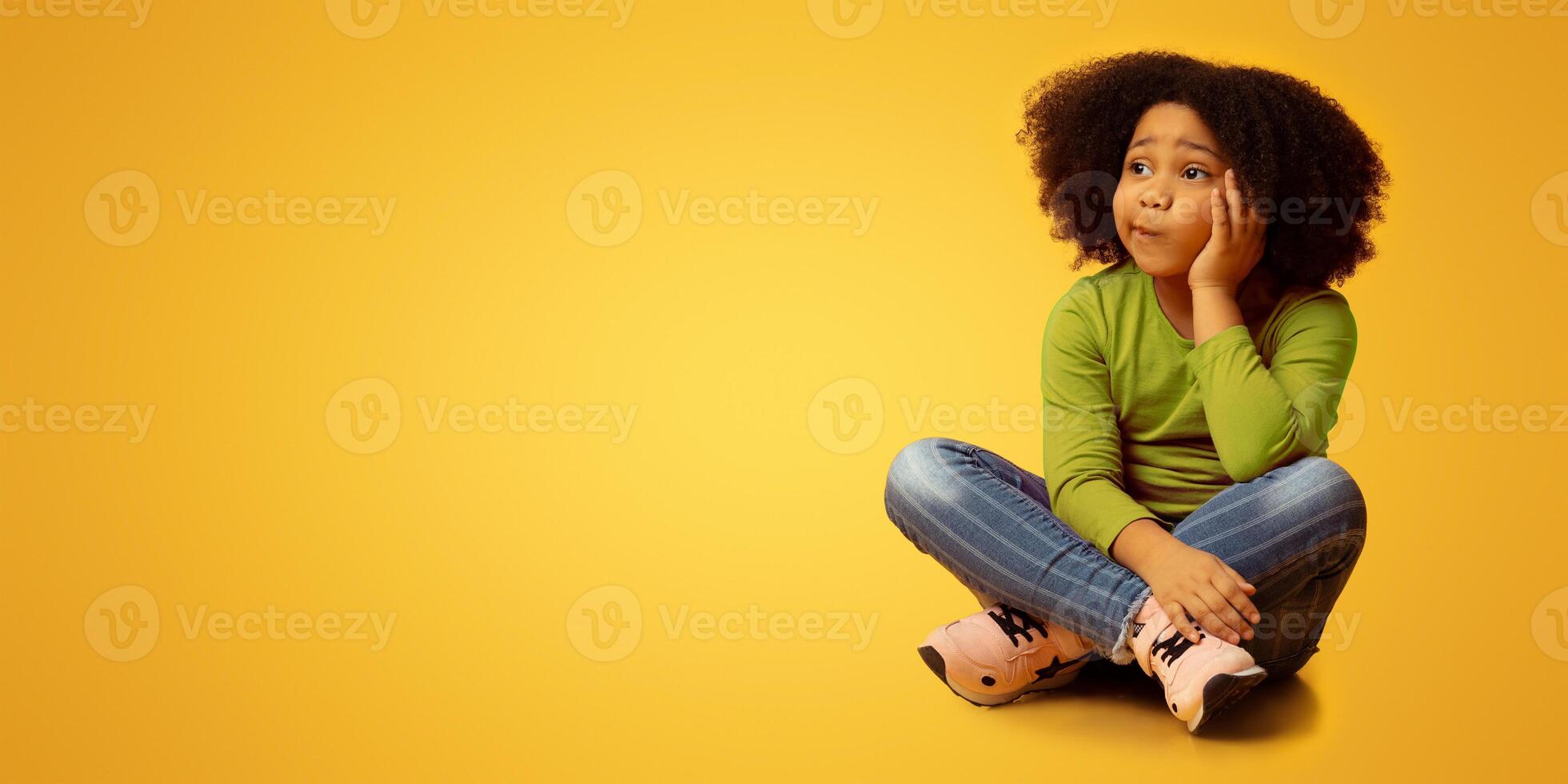 Pensive little black girl sitting on floor over yellow background in studio and thinking, looking away at copy space, panorama photo