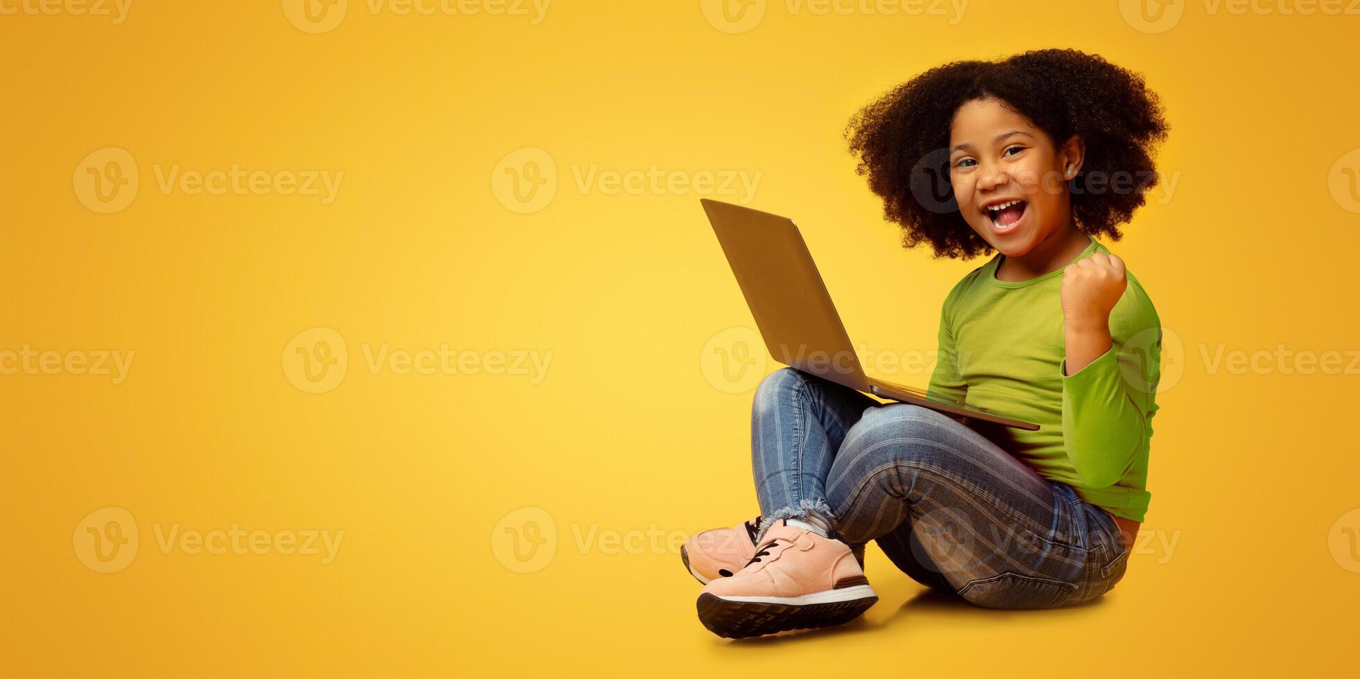 Joyful little african american girl sitting with laptop on lap and celebrating success, raising fist and emotionally shouting on yellow background photo
