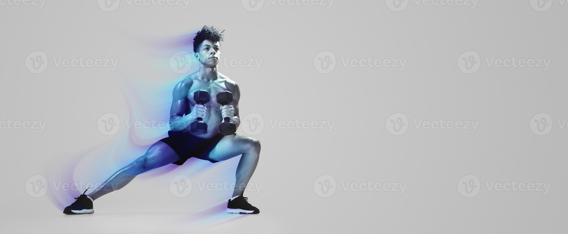 Athletic young man performing deep lunge while holding dumbbells, exercising on gray studio backdrop, full length. Strength and discipline, gym workout with weights concept photo