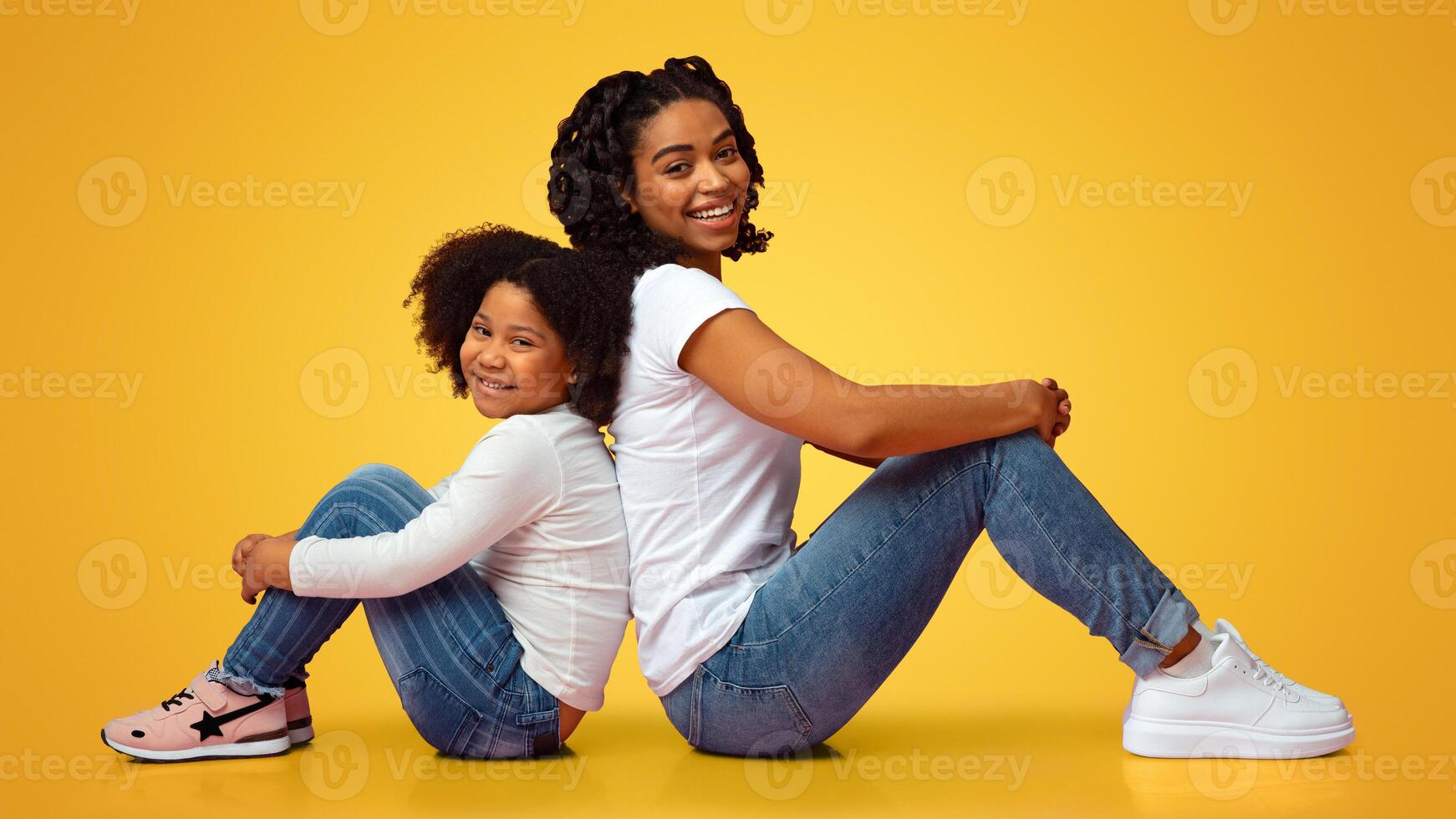 Like Mother Like Daughter. Little African American Girl And Her Mom Sitting Back To Back On Floor In Studio Over Pink Background, Side View photo