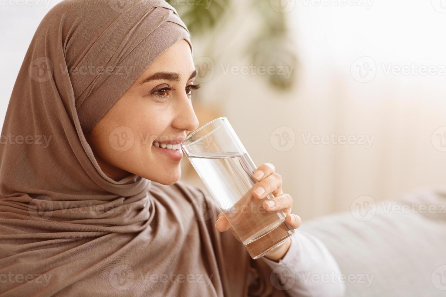 Healthy Liquid. Smiling Muslim Woman In Hijab Drinking Mineral Water From Glass At Home, Closeup Image With Selective Focus, Free Space photo