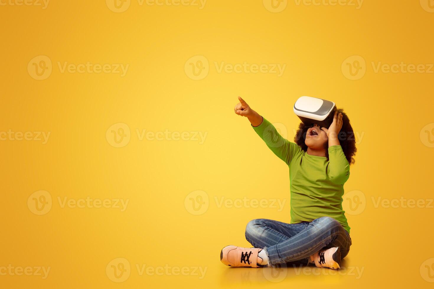 Little Afro Girl Wearing VR Headset, Experiencing Virtual Reality And Pointing Away While Sitting On Floor Over Yellow Background In Studio, Panorama photo