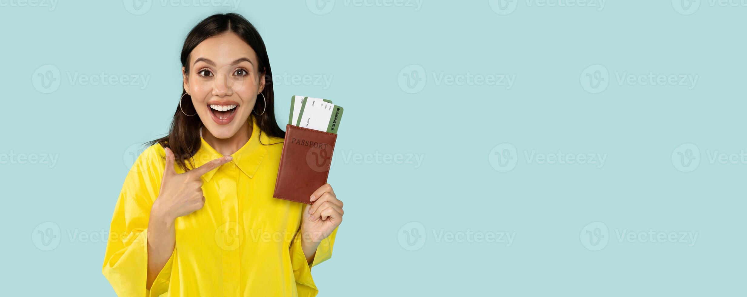 Portrait of happy beautiful brunette young woman wearing yellow shirt pointing at traveling flight tickets and passport isolated over blue studio background. Tourism, cheap tickets, deals photo