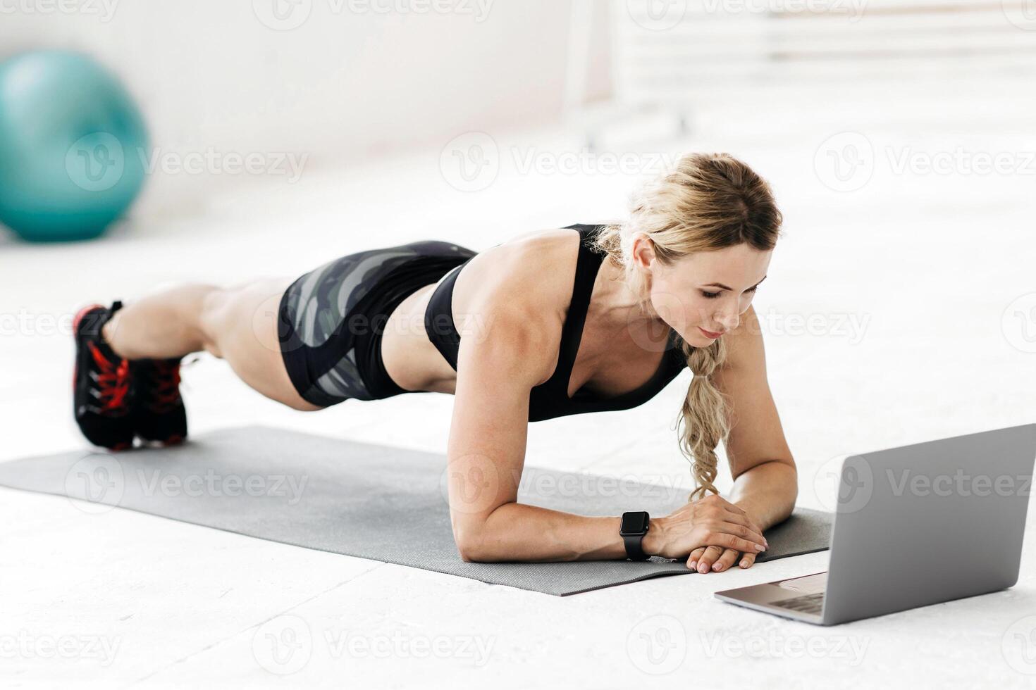 Online lesson at home during coronavirus pandemic. Attractive mature sportswoman in sports uniform makes plank on mat with fitness ball and bottle of water on floor and looks in laptop, free space photo