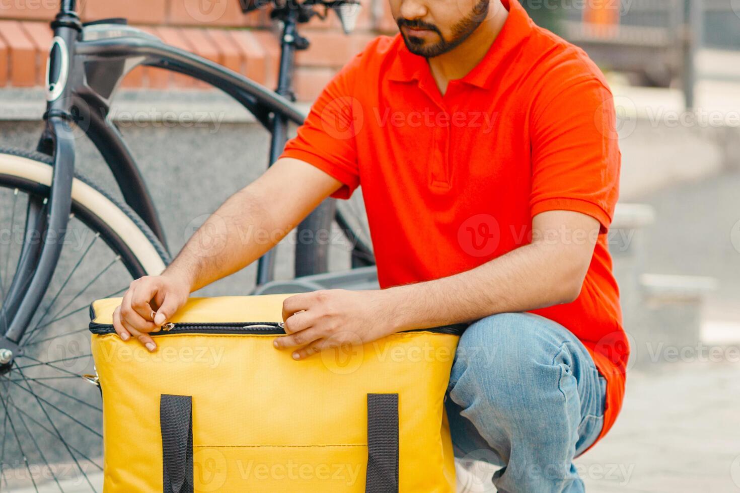Accurate delivery. Busy bearded courier zips up large yellow bag for client, beside a bicycle on street, outside, free space photo