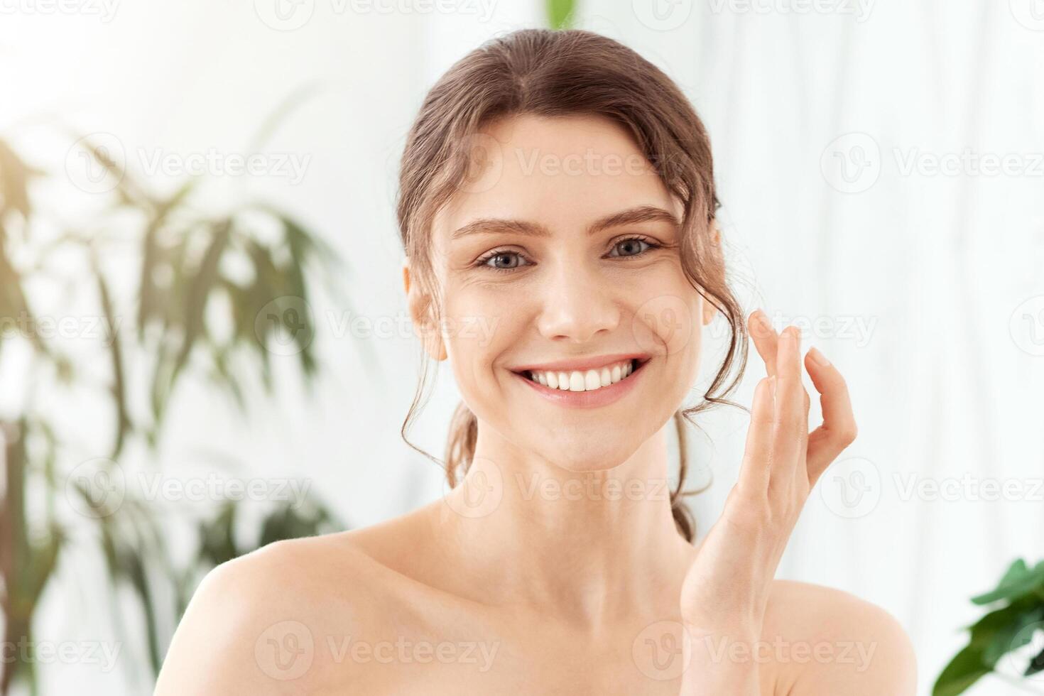 Attractive young woman using face cream at home, looking at camera and smiling, empty space. Beautiful lady with ponytail holding cream jar and applying beauty product on her face, bedroom interior photo