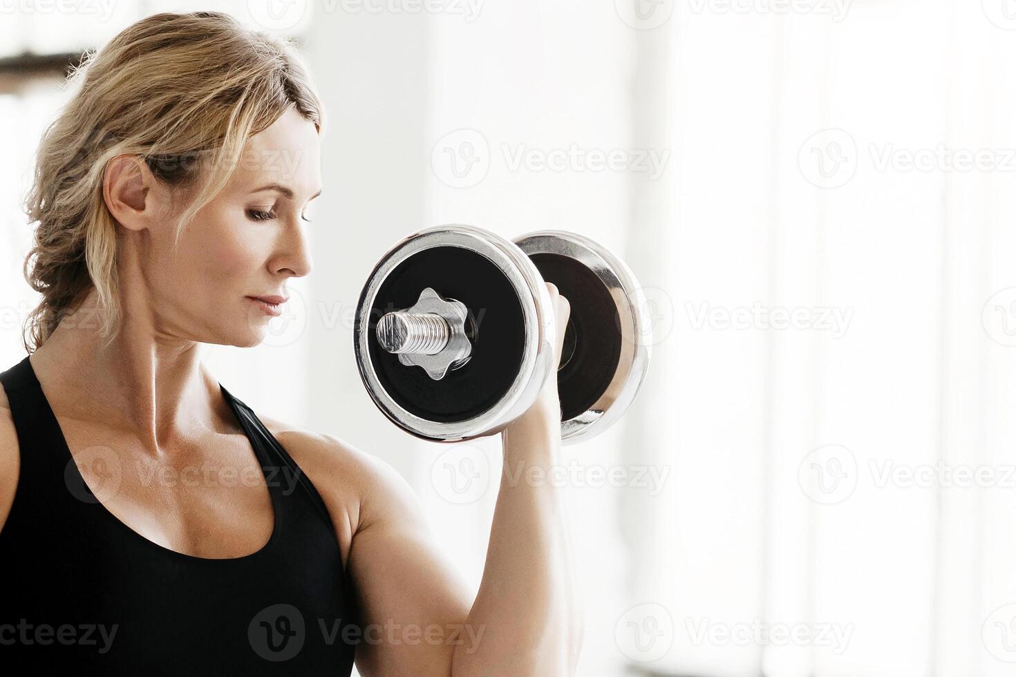 Strong muscular trainer at workout in gym. Adult woman in sportswear with fitness tracker doing exercises with dumbbells at daylight, copy space photo