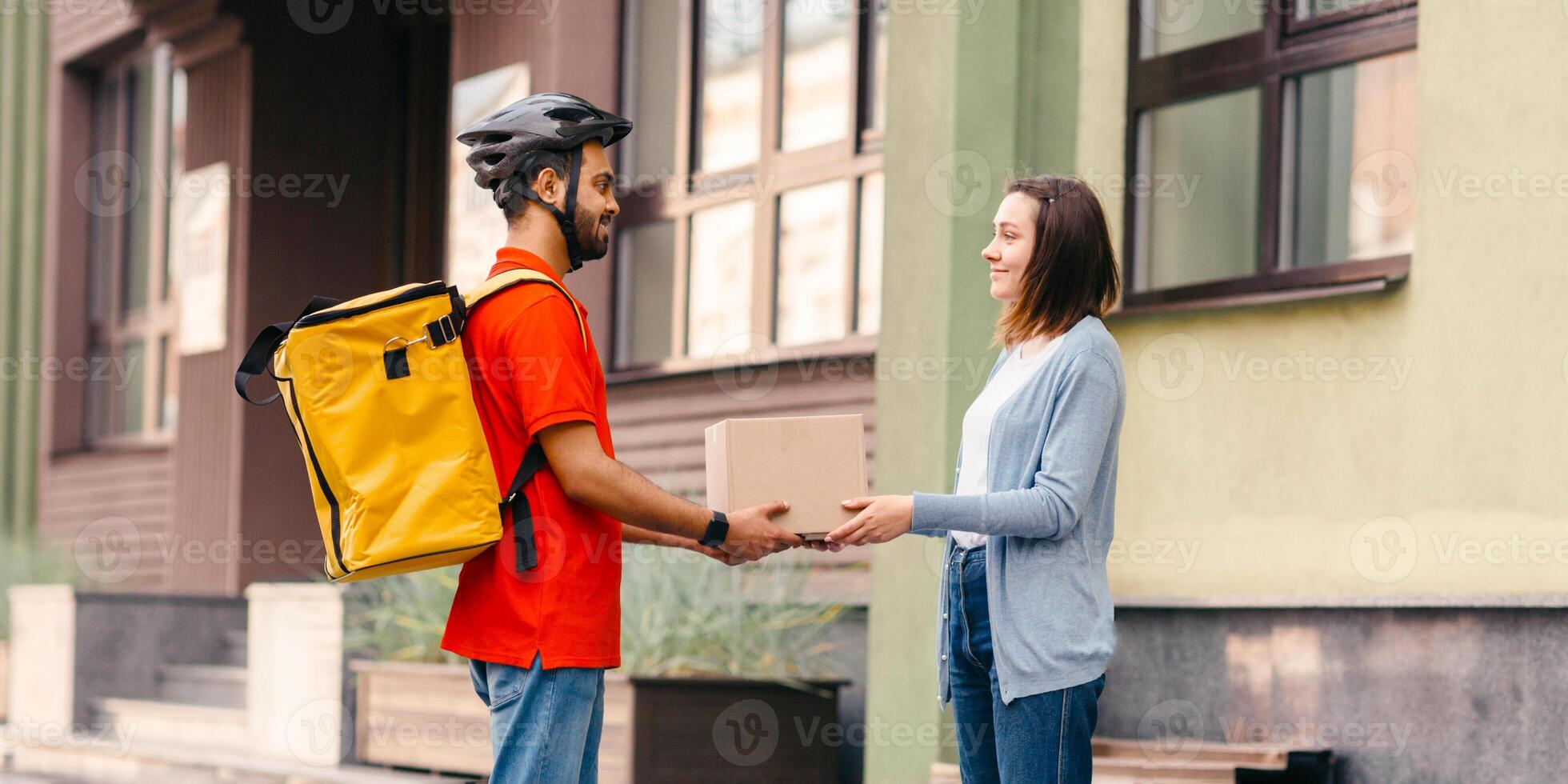 Courier services and online shopping. Young man with yellow backpack, helmet and bicycle gives girl a cardboard box, outdoors, side view, free space photo