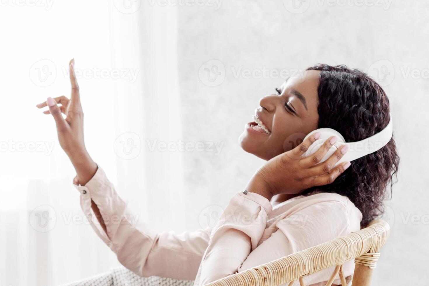 Joyful African Woman Singing While Listening Music With Wireless Headphones At Home, Sitting In Stylish Wicker Chair, Enjoying Favorite Song Playlist, Dancing With Hand, Side View With Copy Space photo