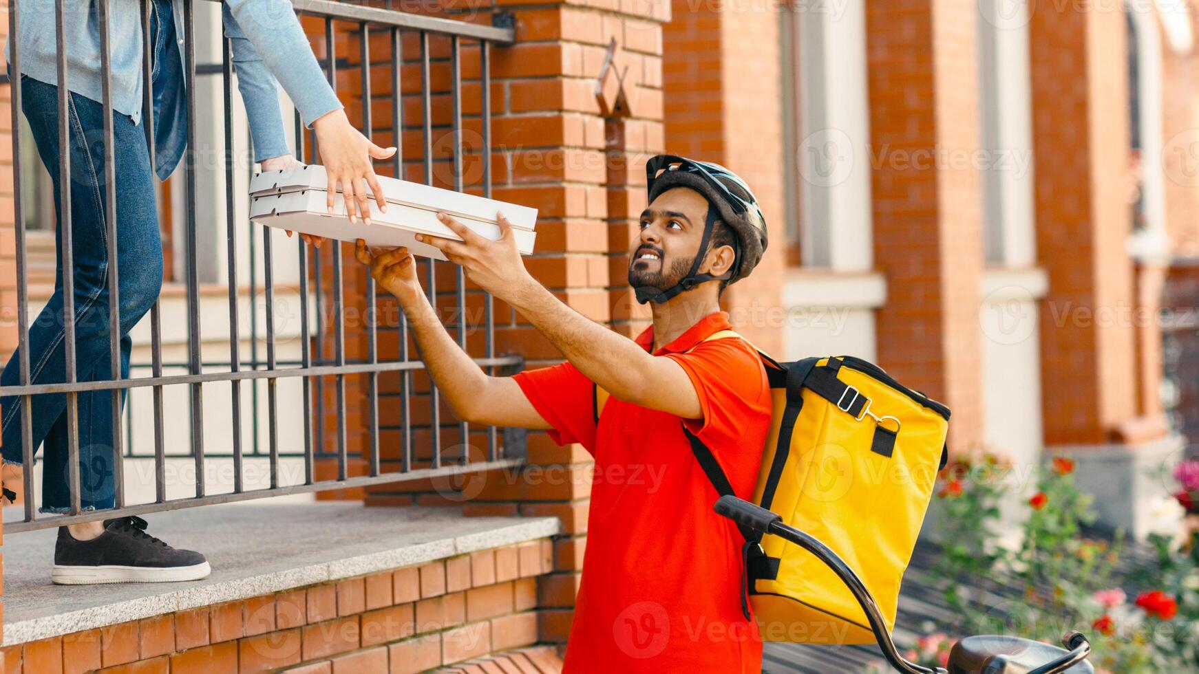 Meeting courier and client. Smiling deliveryman in helmet with big yellow backpack on bicycle delivered pizza to entrance for client, panorama, free space photo