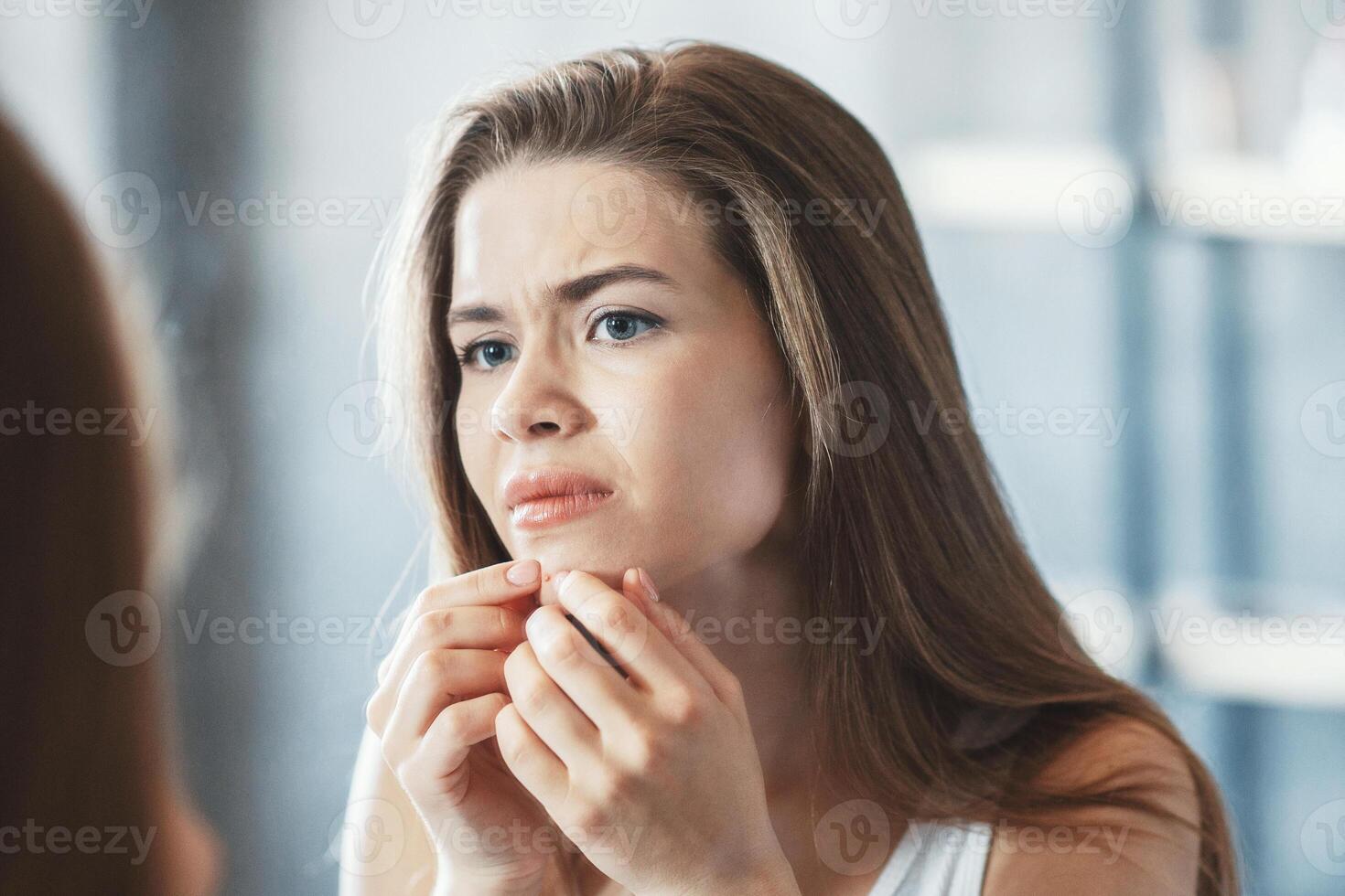 Skin problem. Young girl squeezing her pimple in front of mirror in bathroom photo
