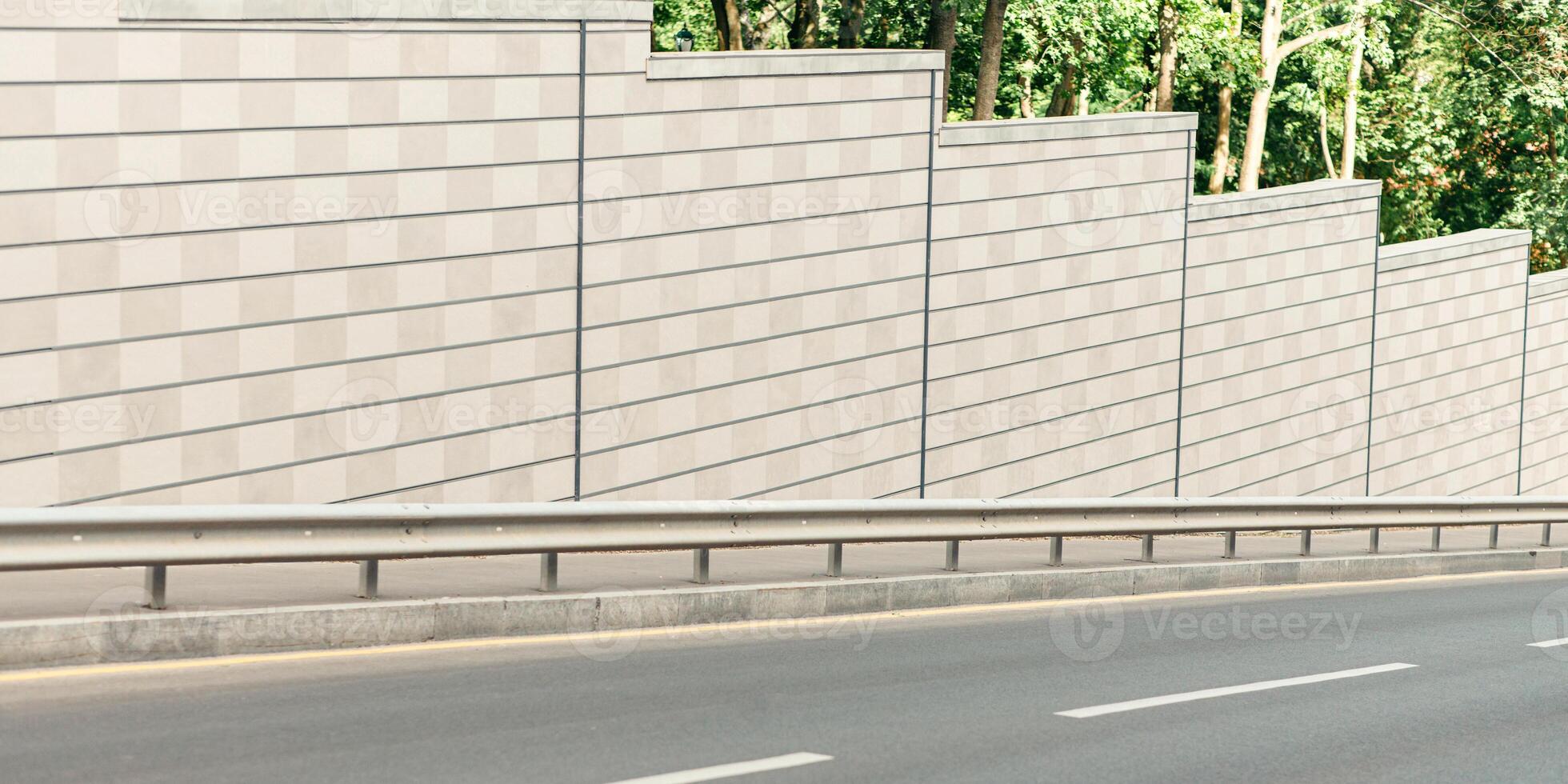 Modern road in city near big green park. Amazing view of empty highway in town street with gray stone walls on sides and with pedestrian sidewalk in summer sunny day, anyone, free space, outdoor photo