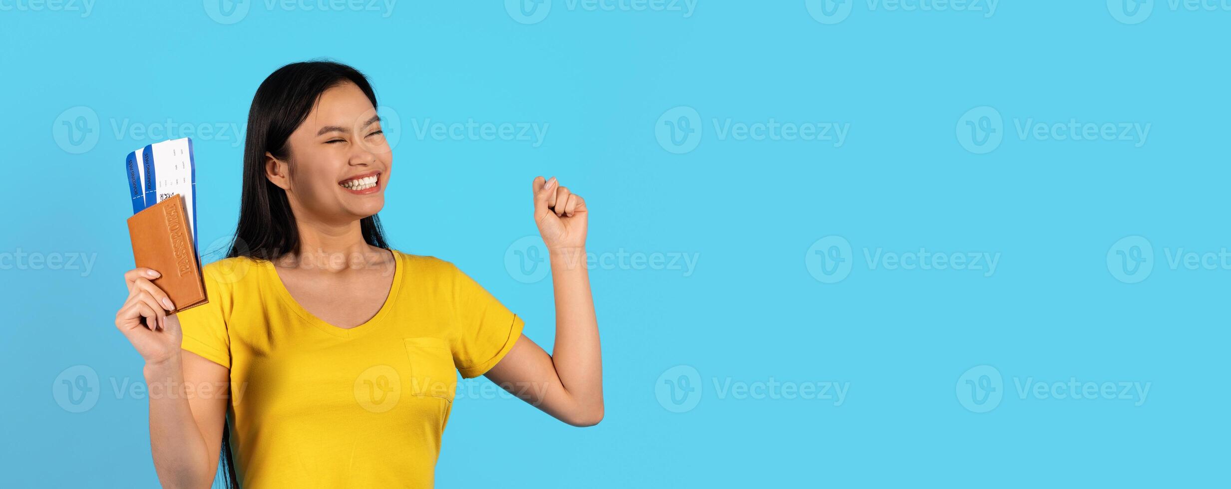 Satisfied excited young chinese woman in yellow t-shirt with passport, air tickets looks at empty space, rejoices to travel, isolated on blue background. Journey, ad and offer and human emotions photo