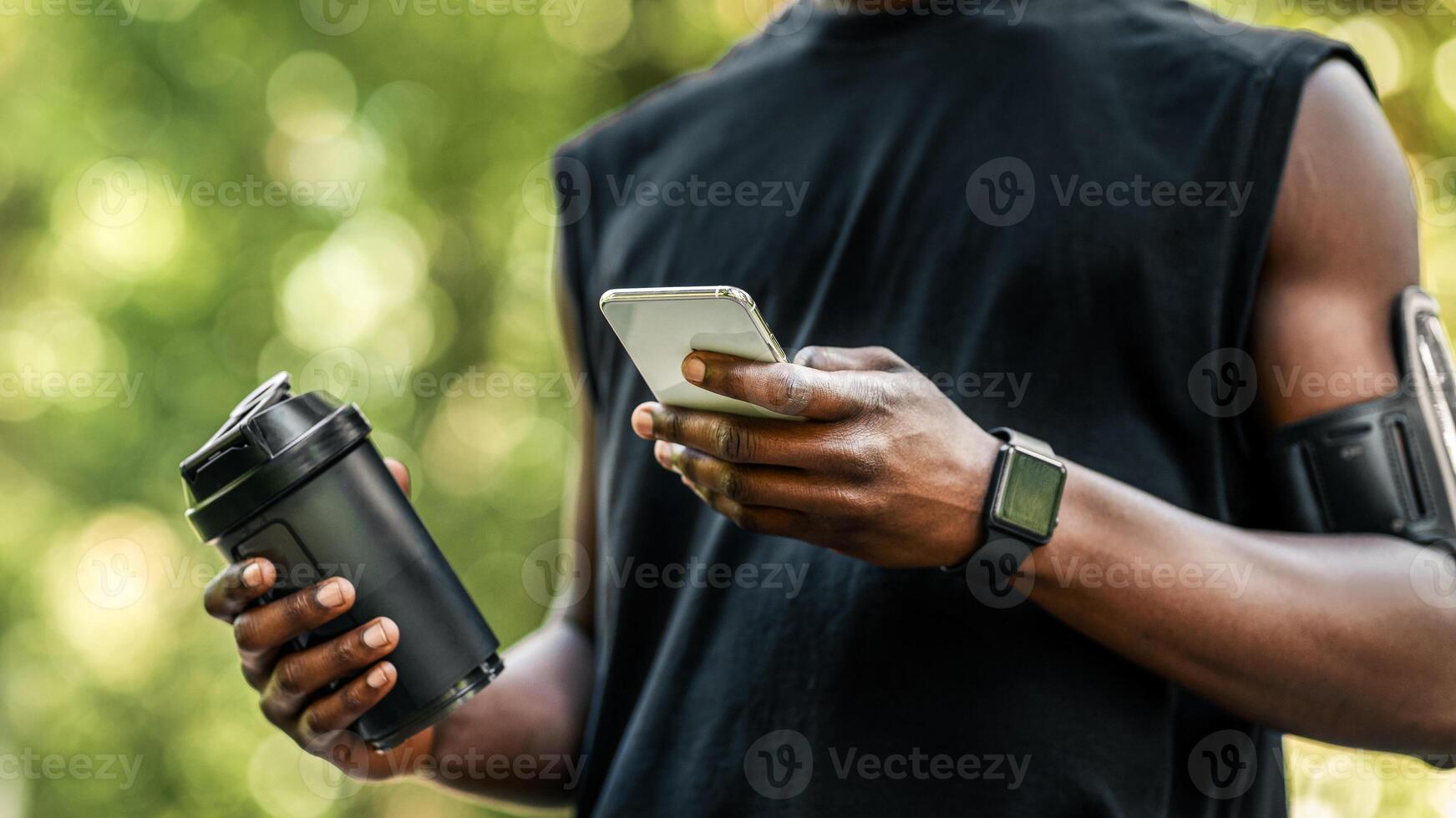 Fitness mobile application. Cropped of african sportsman checking on sport app on mobile phone during break, holding bottle with protein drink, blurred park background, copy space photo