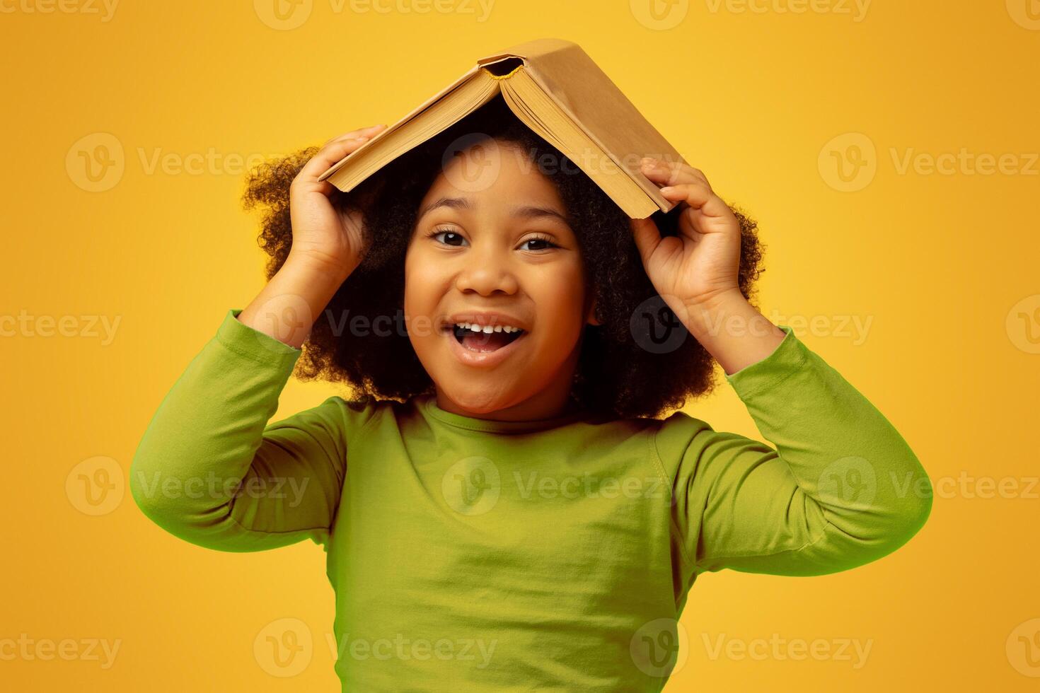 Reading Is Fun. Portrait Of Positive Black Little Girl Holding Opened Book On Her Head, Fooling Over Yellow Background, Free Space photo