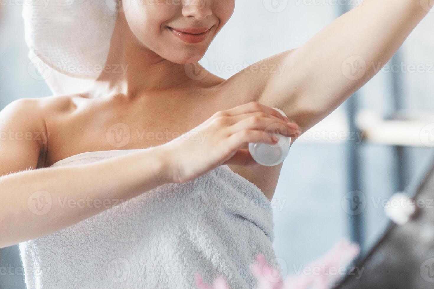 Beautiful young woman applying deodorant on armpit in front of mirror at bathroom photo