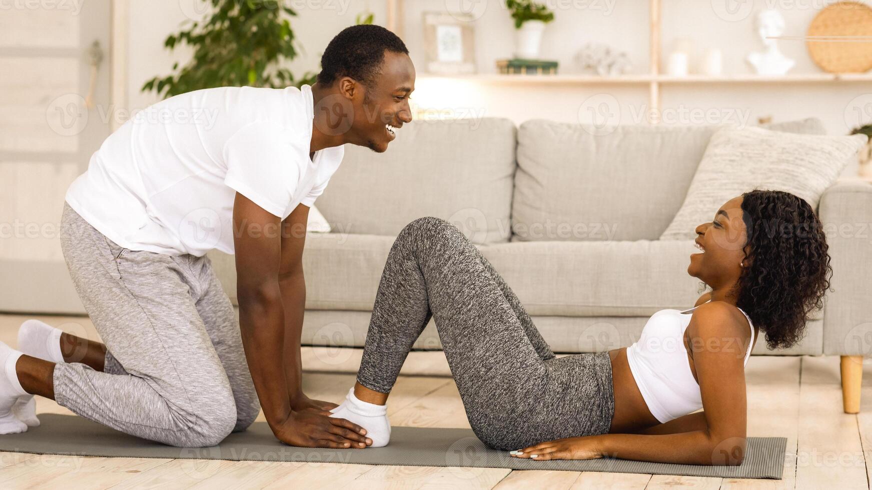 Athletic black couple exercising together at home, working on abs. Cheerful guy in sportswear holding his girlfriend legs while she is doing exercises for belly, having sport together photo