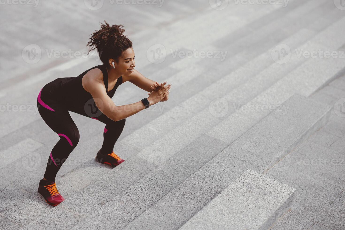 Jumping and squats on stairs. Smiling african american girl with wireless headphones and fitness tracker doing exercises on stairs in city, free space photo