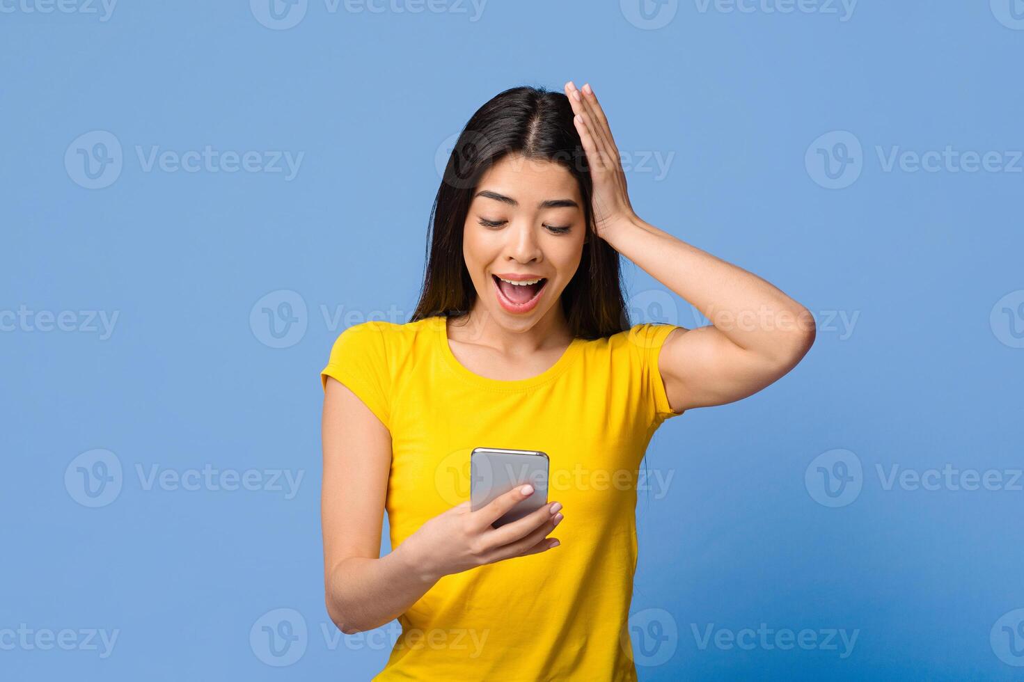 Mobile Offer. Amazed asian woman holding smartphone, looking at device screen with excitement, standing with open mouth over purple studio background, emotionally reacting to great ad, copy space photo