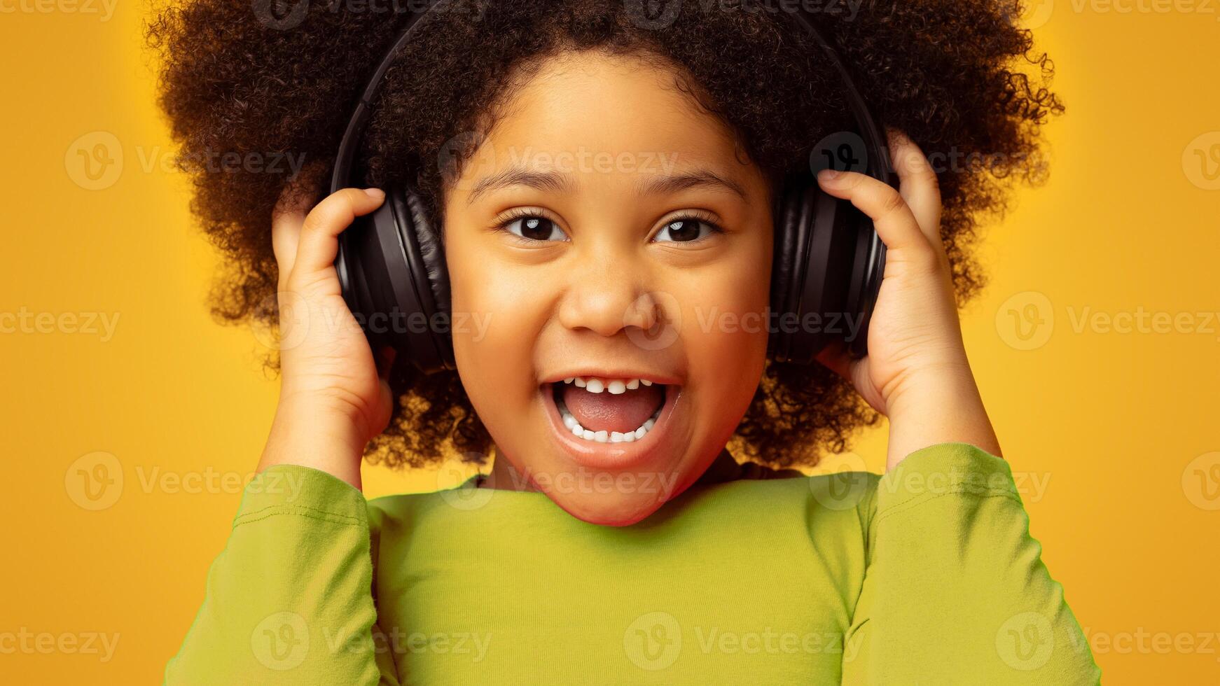 Closeup Portrait Of Excited Little African Girl Enjoying Listening Music In Wireless Headphones, Posing Over Yellow Background And Looking At Camera photo
