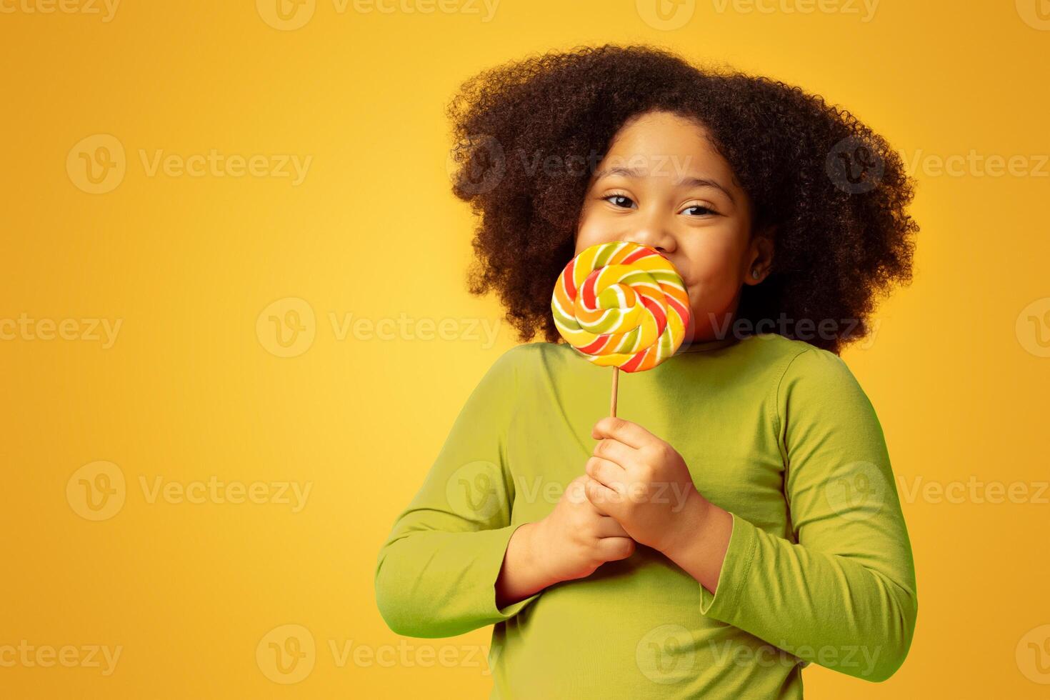 Positive little afro girl licking lollipop candy, enjoying sugar treat and looking at camera, posing over yellow background, panorama with copy space photo