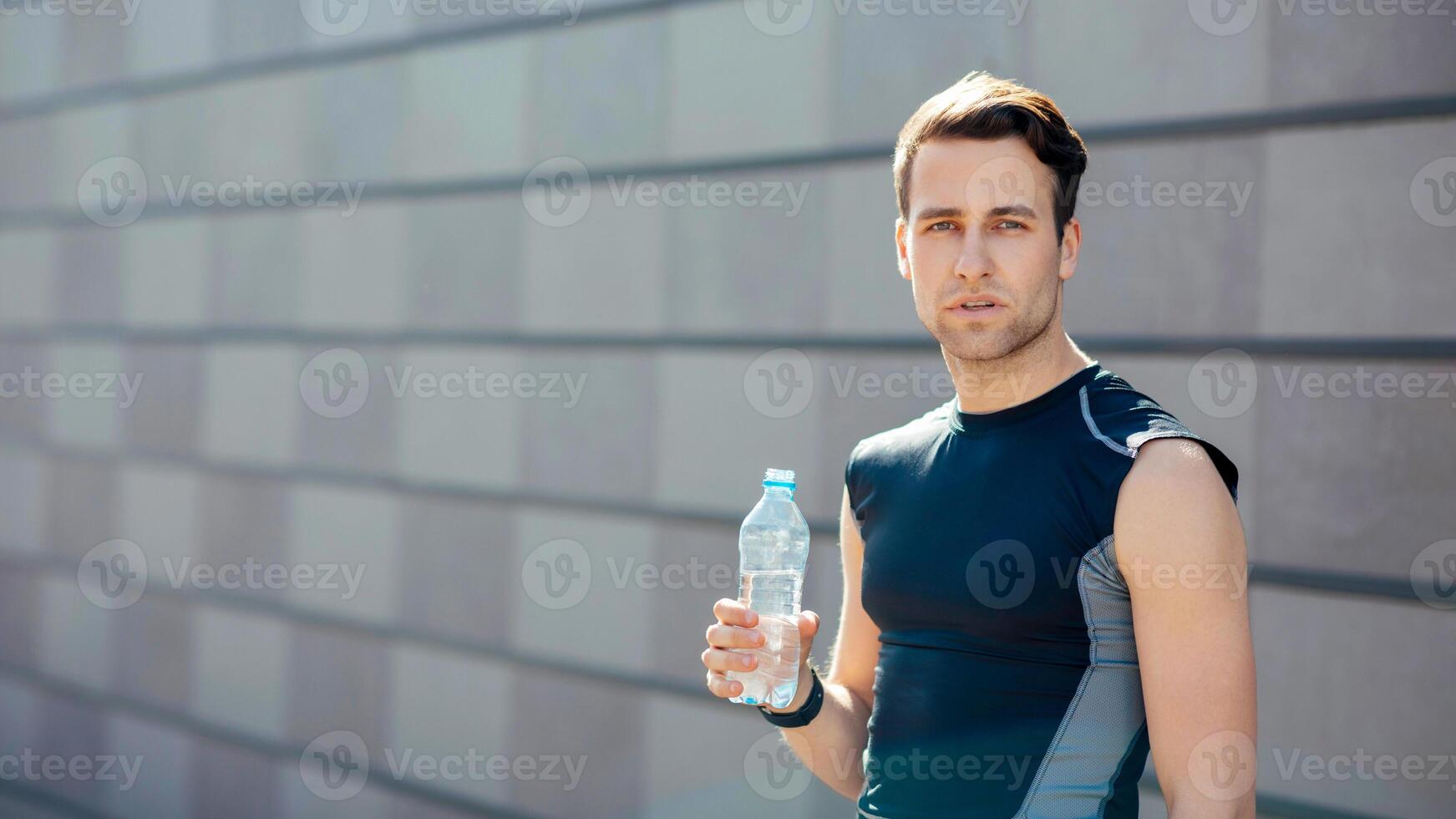 Training, thirst and water. Tired young man in sportswear holds bottle in hand, on gray wall background, free space, outdoor photo