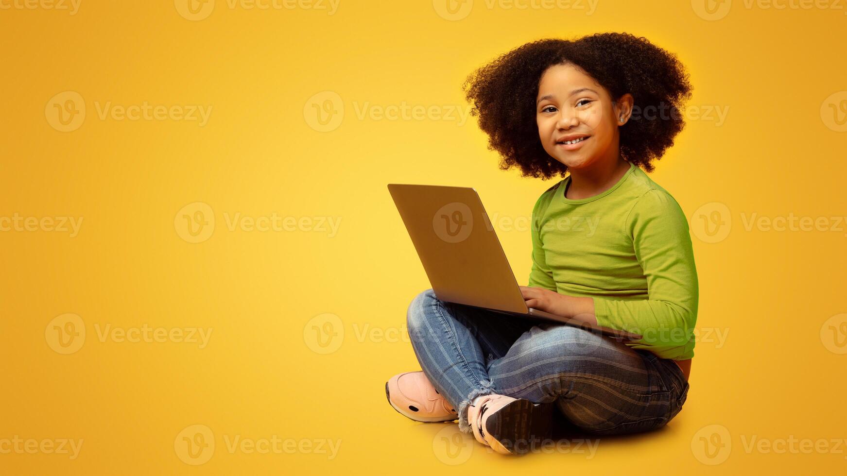 Online Education Concept. Adorable Little Afro Girl Sitting On Floor With Laptop And Looking At Camera, Posing Over Yellow Background With Empty Space photo