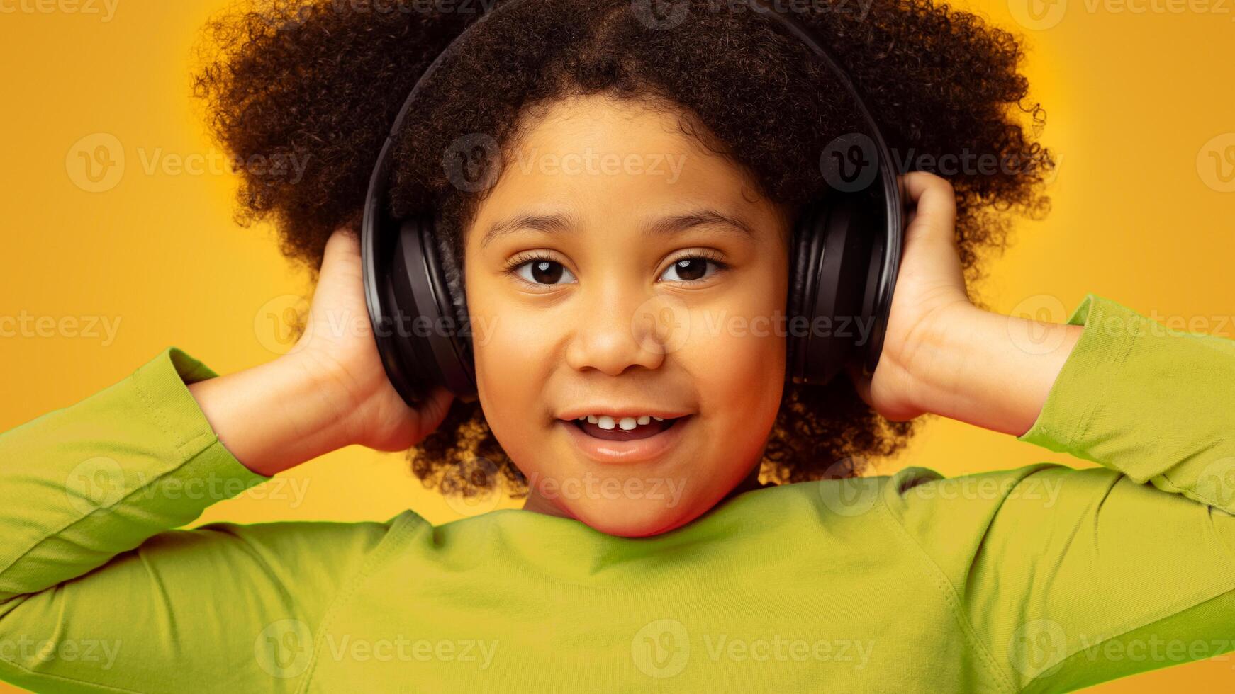 Closeup Portrait Of Cute Little Black Girl In Wireless Headphones Over Yellow Background, Listening Her Favorite Music Online photo