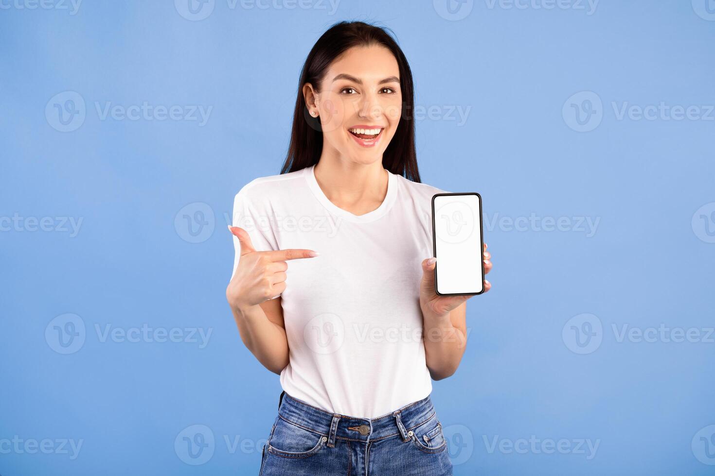 Try This. Excited woman pointing at smart phone with white blank screen for advertisement, blue wall, mock up photo