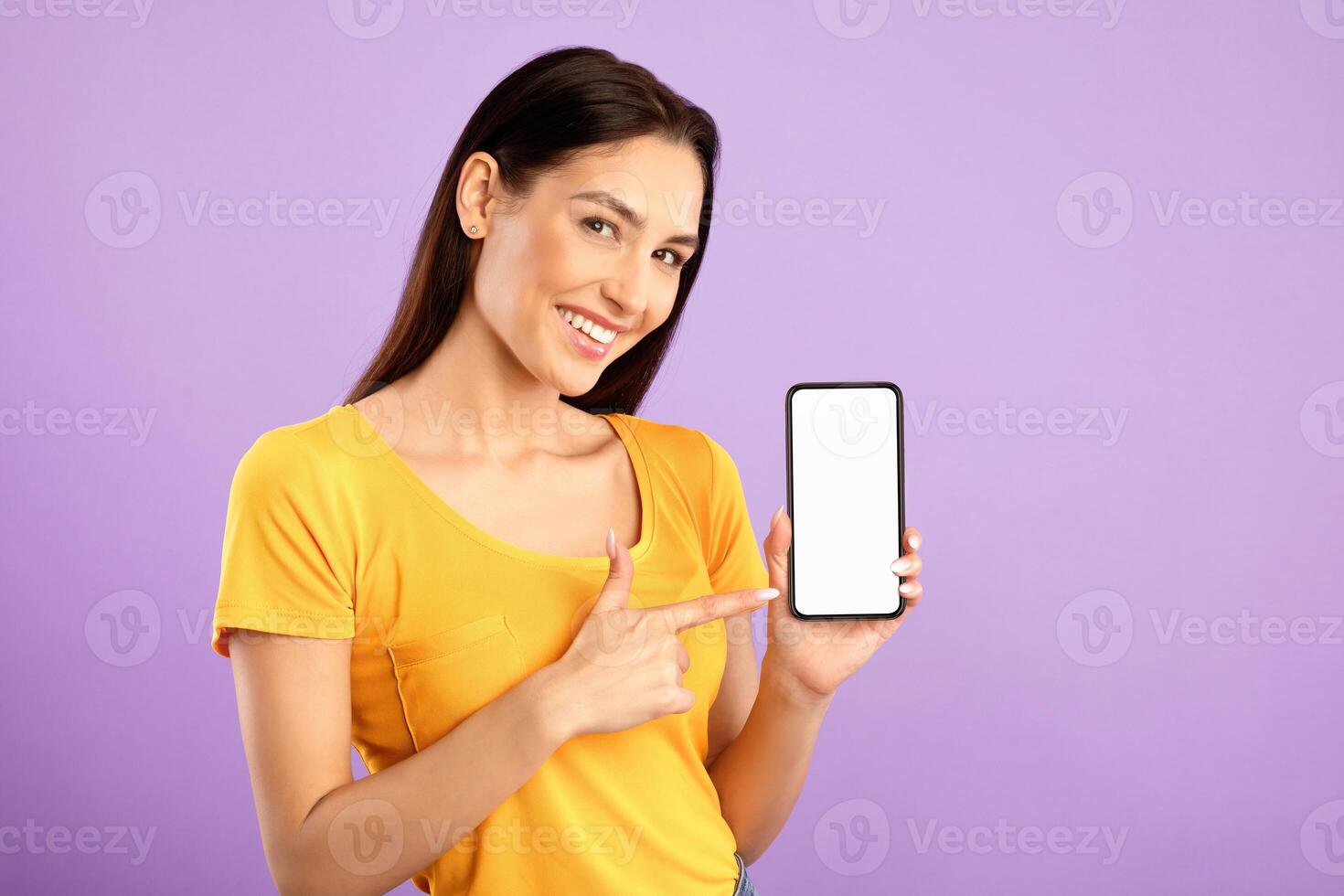 Try Something New. Happy girl pointing at cell phone with white blank screen for advertisement, purple wall, mock up photo