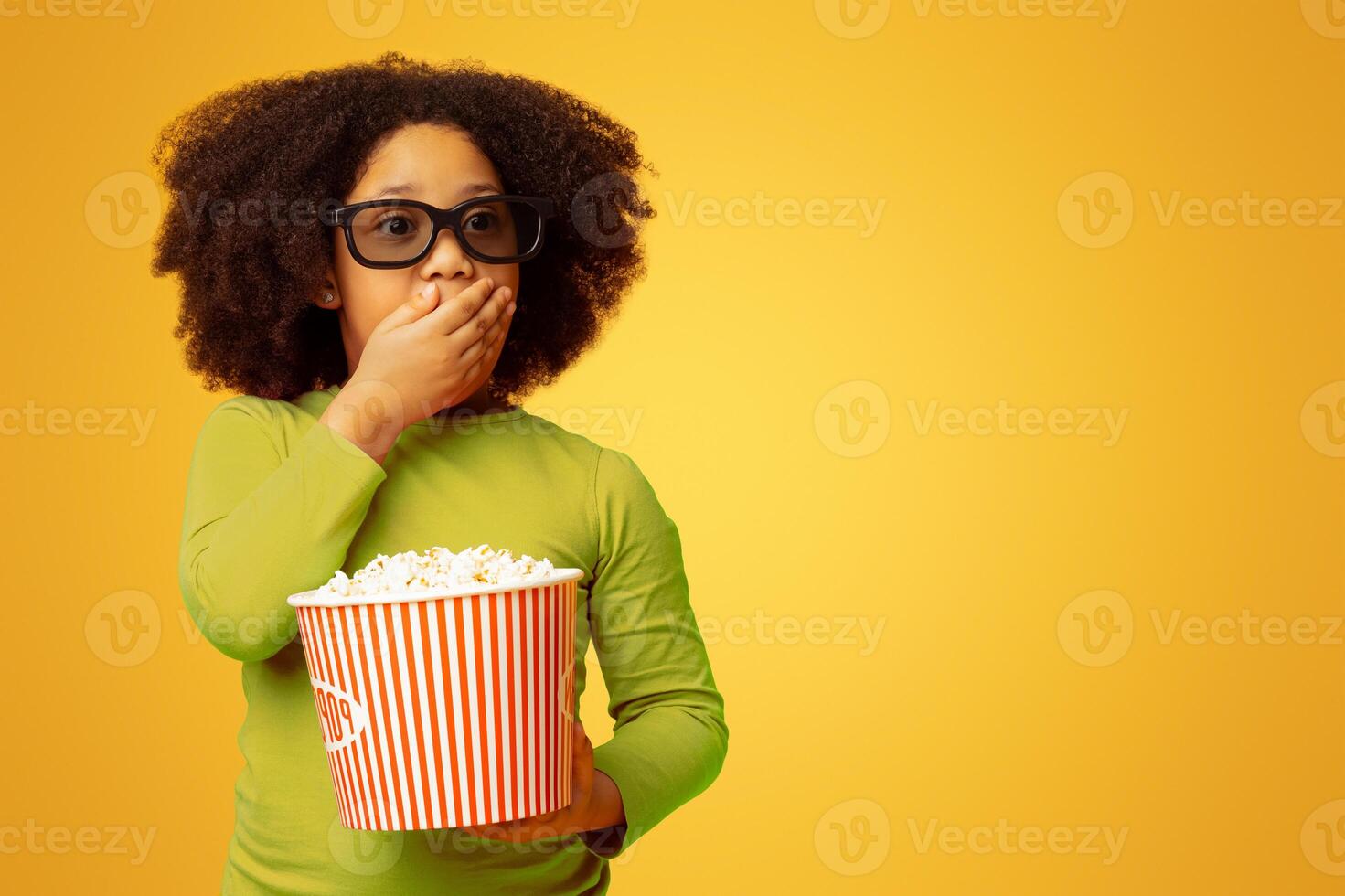 Scary Movie. Shocked little african girl in 3d glasses and with popcorn, covering mouth with hand and looking with frightened face expression photo